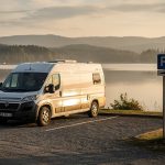 Comment repérer les panneaux qui limitent le stationnement des camping-cars ? Camping-car blanc arrivant sur un parking communal au bord d’un lac, sous une douce lumière de fin de journée.