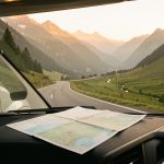 Préparer un road trip en camping-car : 8 règles pour partir serein Camping-car moderne sur une route panoramique avec une carte pliée au premier plan, devant un paysage naturel éclairé par le soleil levant.